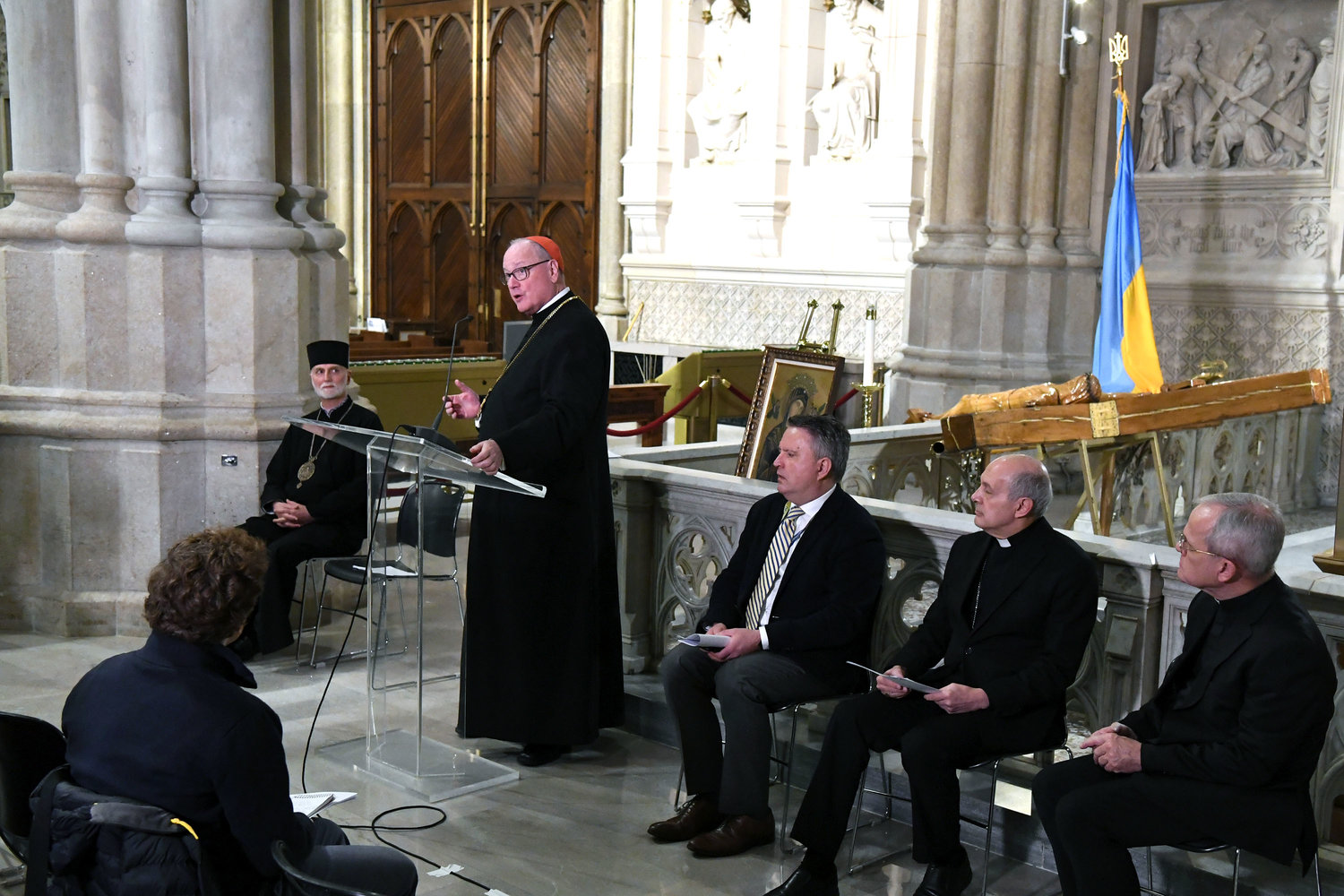 Cardinal Dolan speaks at a March 24 press briefing at St. Patrick's Cathedral about the war in Ukraine and U.S. Church efforts to assist refugees and other displaced persons in Ukraine. Seated, from left, are Archbishop Borys Gudziak of the Ukrainian Catholic Archeparchy of Philadelphia; Ambassador Sergiy Kyslytsya, Ukraine's ambassador to the United Nations; Archbishop Gabriele Cacchia, Vatican nuncio to the United Nations; and Msgr. Peter Vaccari, president of Catholic Near East Welfare Association (CNEWA), a papal agency headquartered in Manhattan. In the background is the Ukrainian Cross of Gratitude, positioned next to the Ukrainian flag.